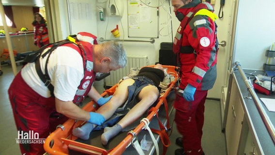 Sanitätspersonal bei einer Seenotrettungsübung versorgen eine Person auf einer Trage im Innenraum eines Schiffs. © Screenshot 