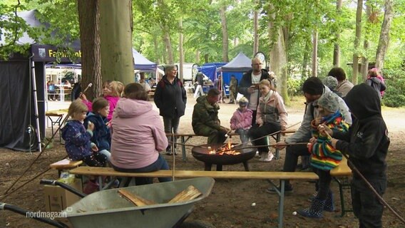 Mehrere Familien mit kleinen Kinder sitzen um eine Feuerschale herum in einem waldigen Gelände und grillen Marshmallows an Stöckern. © Screenshot 