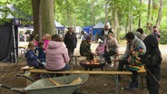 Mehrere Familien mit kleinen Kinder sitzen um eine Feuerschale herum in einem waldigen Gelände und grillen Marshmallows an Stöckern. © Screenshot 