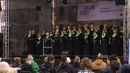 Ein Chor mit Schwarzen Roben und grünen Halskrausen tritt beim Chorfest in Demmin auf einer Freilichtbühne vor Publikum auf. © Screenshot 