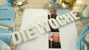 Auf einer hälzernen Tischplatte liegen die Buchstaben der Wörter "Die Woche" nebst mehreren kleinen Utensilien wie Passbildern, Stiften und einem Gebäckstück. © Screenshot 