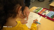 Eine Hand zeigt einem Grundschulkind auf einem Arbeitsblatt mit dem Buchstaben A etwas. © Screenshot 