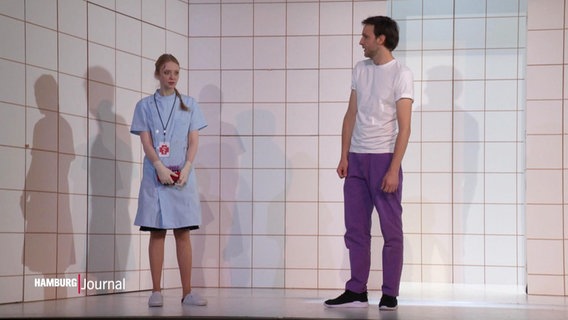Zwei Schauspielende stehen vor einem Bühnbild aus Kachelwänden. Er trägt Alltagsklamotten mit einer lila Hose, sie trägt einen kurzärmeligen Laorkittel. © Screenshot 