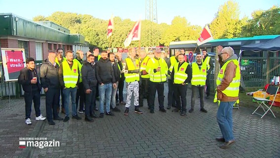 Eine Gruppe streikender Omnibusfahrer steht mit Warnwesten als Pulk auf einem Betriebsgelände. © Screenshot 