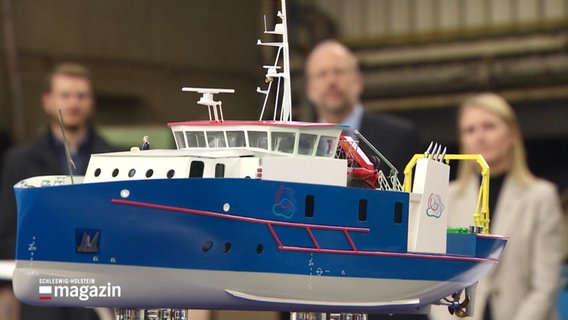 Ein Modell eines kleineren Schiffs steht auf einem Sockel vor drei im Hintergrund unscharfen Personen. © Screenshot 
