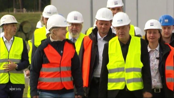 Bundeswirtschaftsminister Robert Habeck (Bündnis 90/Die Grünen) geht mit einer Truppe Begleiter in Warnweste und mit Schutzhelm auf dem Gelände des LNG-Terminals in Lubmin. © Screenshot 