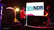 Auf einer Bühne wird ein Schild, das 30 Jahre NDR in MV feiert, getragen © Screenshot 