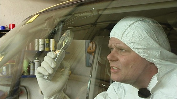 Ein Mann sitzt in Schutzkleidung in einem Auto und untersucht eine Scheibe mit einer Lupe. © Screenshot 