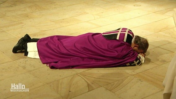 Bischof Bode liegt in einer lila Robe mit verschränkten Armen auf dem Bauch in einer Kirche auf dem Boden. © Screenshot 