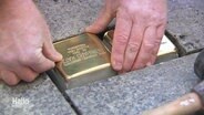 Zwei Hände setzt einen gold-glänzenden Stolperstein neben einen anderen in den Gehweg ein. © Screenshot 