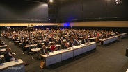 In einem großen Konferenzsaal sitzen viele Menschen in Anzügen an langen Tischreihen nebeneinander. © Screenshot 