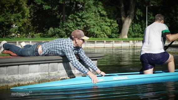 Ein Mann mit Cappie hält vom Steg aus ein Olypia-Kanu im Wasser aufrecht in dem ein weiterer Mann mit Paddel kniet und balanciert. © Screenshot 