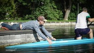 Ein Mann mit Cappie hält vom Steg aus ein Olypia-Kanu im Wasser aufrecht in dem ein weiterer Mann mit Paddel kniet und balanciert. © Screenshot 
