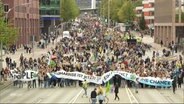 Auf einer mehrspurigen, großen Straße hat sich ein Demozug der Fridays For Future Bewegung positioniert, der ein großes Banner in die Luft hält. © Screenshot 