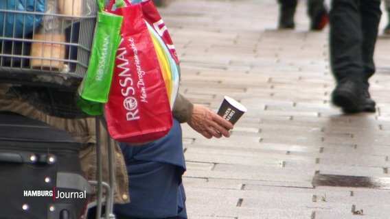 Eine Hand mit einem Kaffeebecher hinter einem vollbepackten Einkaufswagen. © Screenshot 