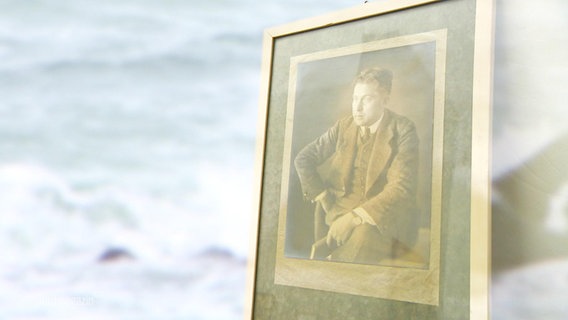 Ein altes Foto in einem Bilderrahmen auf dem ein Mann abgebildet ist, im Hintergrund das Meer. © Screenshot 