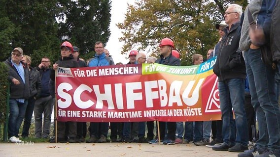 Werftmitarbeitenden stehen hinter einem großen Transparent mit der Aufschrift: " Schiffbau - Für eine starke maritime Industrie an der Küste". © Screenshot 