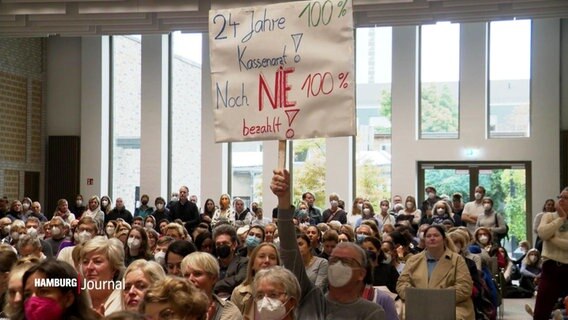 Protestierende Mediziner sitzen in einem Saal, einer hält ein Schild in die Höhe. © Screenshot 