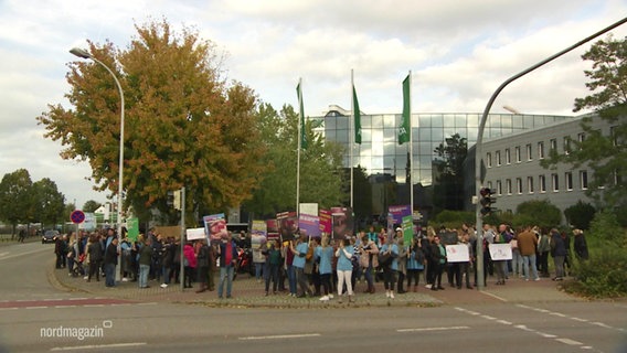 Eine Gruppe von Menschen hält Protestschilder hoch. Darunter auch Menschen, die Arbeitskleidung aus einem Krankenhaus tragen. © Screenshot 