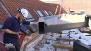 Ein Bauarbeiter mit einem großen Bauteil auf einem Dach. © Screenshot 