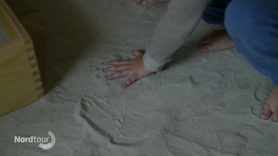 Die Hand und der Fuß eines Kindes in einem Sand-Spiel-Raum in Petersdorf. © Screenshot 