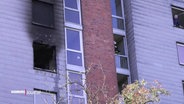 Blick auf ein Fenster eines mehrstöckigen Wohnhauses. Links ist ein Fenster ohne Scheiben, die Ränder der Wand drumherum sind von Flammen schwarz gefärbt. © Screenshot 