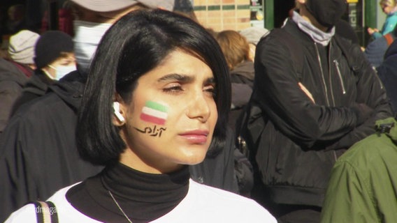 Eine Frau steht auf einer Demo. Sie hat die iranische Flagge auf ihre rechte Wange gemalt. © Screenshot 