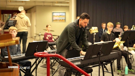 Ein Mann spielt an einem E-Piano in einem Musikraum, um ihn herum sitzen Mitwirkende einer Schul-BigBand. © Screenshot 