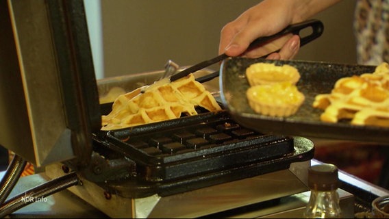 Eine Hand hebt mit einer Zange eine frisch gebackene Waffel aus einem Waffeleisen. © Screenshot 