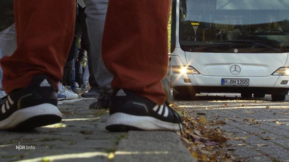 Die Beine von Schulkindern stehen an einer Bushaltestelle an der ein Bus anfährt. © Screenshot 