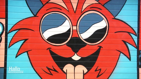 An einer Wand ist eine Cartoon-Katze mit herausgestreckter Zunge und Rundbrille aufgemalt. © Screenshot 