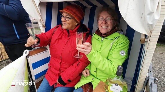 Zwei Frauen mit Sektgläsern in der Hand sitzen lachend in einem Strandkorb © Screenshot 