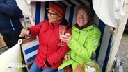 Zwei Frauen mit Sektgläsern in der Hand sitzen lachend in einem Strandkorb © Screenshot 