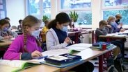 Schülerinnen und Schüler einer Grundschulklasse sitzen an Tischen und tragen Masken. © Screenshot 