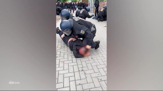 Ein in Schutzuniform bekleideter Polizist kniet sich auf den Nacken eines am Boden liegenden Fußballfans. © Screenshot 