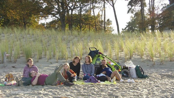 Mehrere Menschen sitzen vor den Dünen am Strand in der Sonne. © Screenshot 