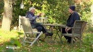 Zwei Männer sitzen in einem Garten an einem Tisch und unterhalten sich. © Screenshot 