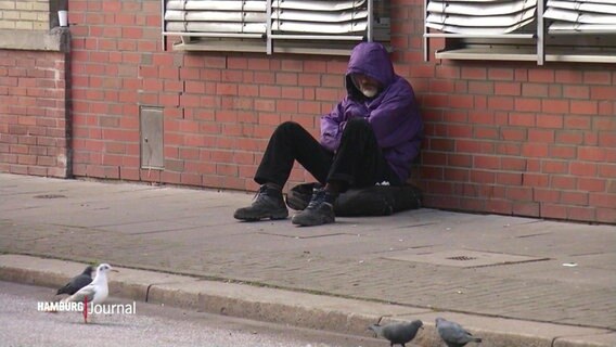 Ein obdachloser Mensch sitzt an eine Häuserwand gelehnt auf der Straße. Die Person hat trägt eine lila Jacke, die Kapuze tief ins Gesicht gezogen. © Screenshot 