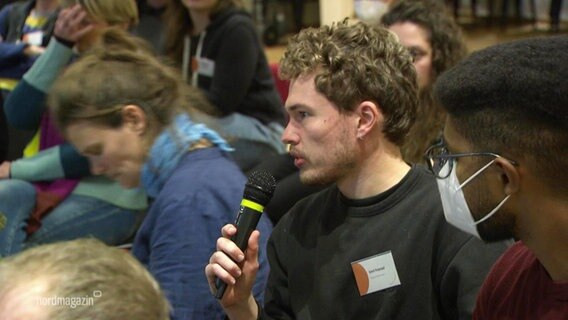 Bei einer Diskussionsrunde spricht ein Teilnehmer aus dem Publikum in ein Mikrofon. © Screenshot 