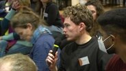 Bei einer Diskussionsrunde spricht ein Teilnehmer aus dem Publikum in ein Mikrofon. © Screenshot 
