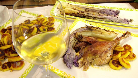 Eine Entenkeule mit Bratkartoffeln und Chinakohl, angerichtet auf einem Teller. Dazu ein Glas Wein. © Screenshot 