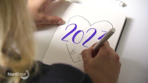 Eine Frau schreibt in ein Heft in schnörkeliger Schrift "2023". © Screenshot 