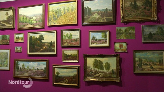 An einer lila Wand hängen mehrere Gemälde der Lüneburger Heide. © Screenshot 