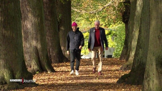 Ein älterer Herr und ein junger Student gehen durch eine herbstliche Allee mit Blätterlaub auf dem Boden. © Screenshot 