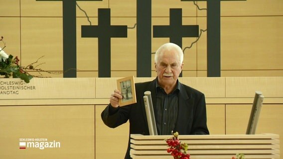 Ein älterer Mann steht im Landtag vor schwarzen Kreuzen und hält ein Bild seines Vaters in die Höhe. © Screenshot 