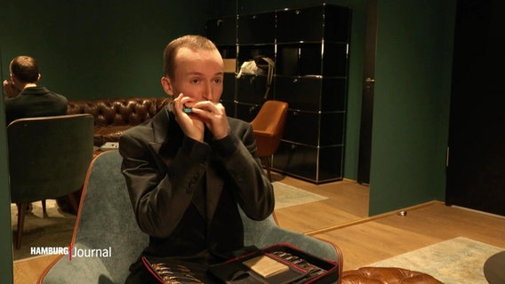 Der Mundharmonikerspieler Konstantin Reinfeld an seinem Paradeinstrument auf einem Stuhl sitzend und in die Kamera blickend. © Screenshot 