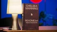Ein Buch von Chelsea Manning steht auf einem Tisch neben einer Lampe. © Screenshot 