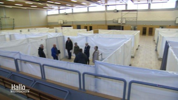 Mehrere Menschen stehen in einer zu einer Flüchtlingsunterkunft umgebauten Turnhalle. © Screenshot 