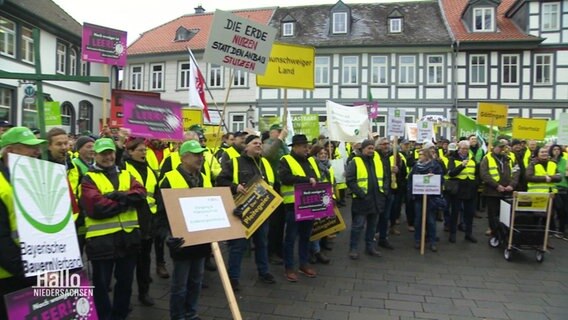 Viele protestierende Landwirte stehen mit gelben Westen und Schildern im Halbkreis in einer Innenstadt. © Screenshot 
