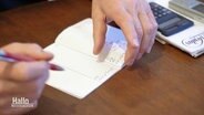 Nahaufnahme von zwei Händen, die etwas in ein Kassenbuch reinschreiben. Daneben liegt ein Taschenrechner. © Screenshot 